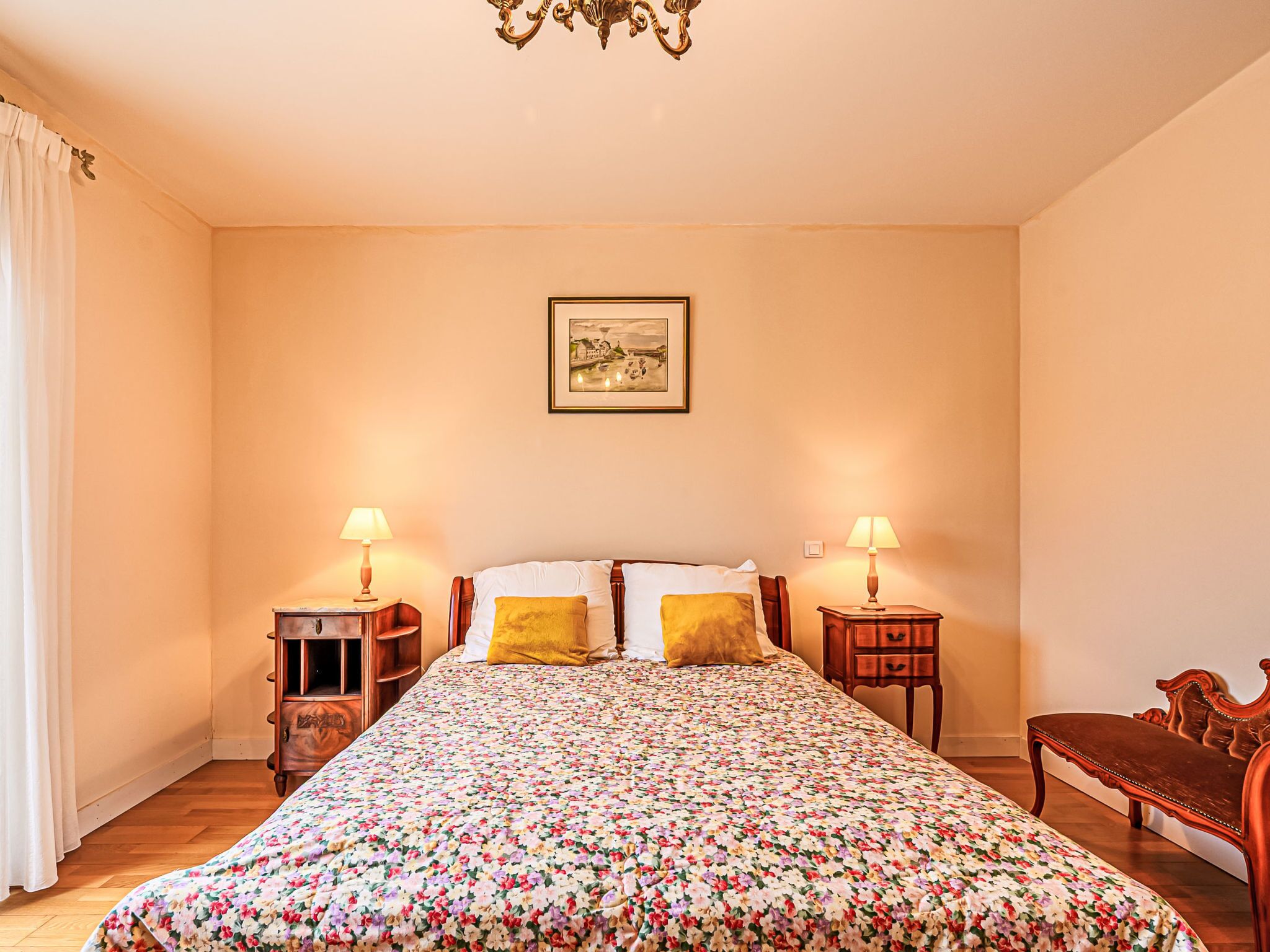 Ein Schlafzimmer mit floralem Bettbezug, zwei Nachttischlampen, einer Holzbank und einem gerahmten Bild an der Wand.