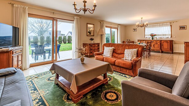 A spacious living room with a sofa, armchairs, a coffee table, and a TV. There is a dining area with a table and chairs, and a kitchen in the background. - Le Prat by Interhome (Guidel)