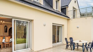 A house with a patio area featuring a dining table and chairs, a sliding glass door leading to a garden, and a second door with a view of the garden. - Le Prat by Interhome (Guidel)