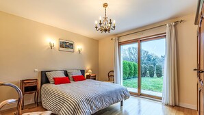 A bedroom with a large bed, wooden flooring, a chandelier, a view of the garden through a sliding door, and a small bedside table with a lamp. - Le Prat by Interhome (Guidel)