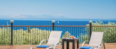 Two lounge chairs with cushions, a small table with a fruit bowl, and a view of the sea and mountains.