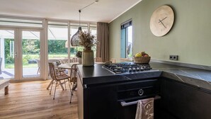 A modern kitchen with a large island, a dining area with wicker chairs, and a view of a garden through glass doors. - 6 pers. Veenhuis Comfort Plus by Interhome (Vlagtwedde)