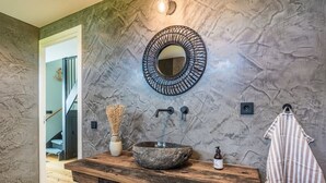 A bathroom with a stone wall, a round mirror, a wooden vanity, a sink, a towel, and a staircase leading to another room. - 6 pers. Veenhuis Comfort Plus by Interhome (Vlagtwedde)