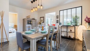 Un espace de repas moderne, avec une table en bois dressée pour quatre personnes, entourée de chaises. On aperçoit une cuisine à l’arrière-plan, et une grande fenêtre offre une vue sur la verdure.
