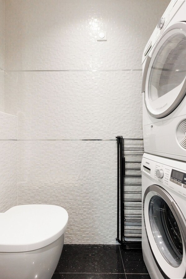 A compact laundry room with a white tiled wall, a black tiled floor, a white toilet, and a front-loading washing machine.