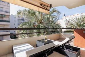A balcony with a sofa, a small table with a bottle, and a view of palm trees and buildings.