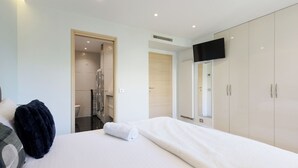 Un dormitorio moderno con cama, armario, televisor y un baño visible a través de una puerta abierta.