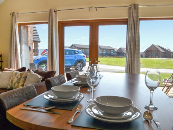 Un coin repas avec une table dressée pour deux personnes, et une vue sur une voiture bleue et des maisons à travers les fenêtres.