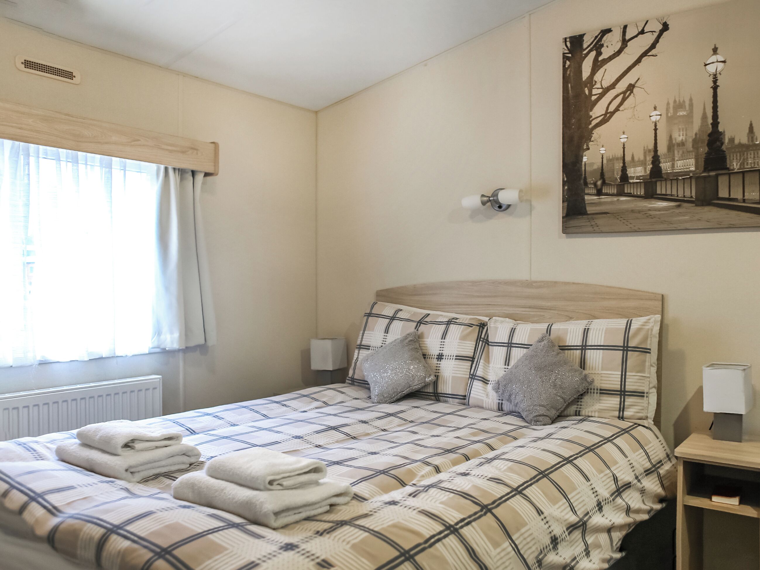 A bedroom with a bed, pillows, a bedside table, a lamp, a radiator, a window with curtains, and a painting on the wall.