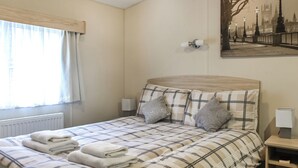 Une chambre à coucher avec un lit, des oreillers, une table de chevet, une lampe, un radiateur, une fenêtre avec des rideaux et un tableau accroché au mur.