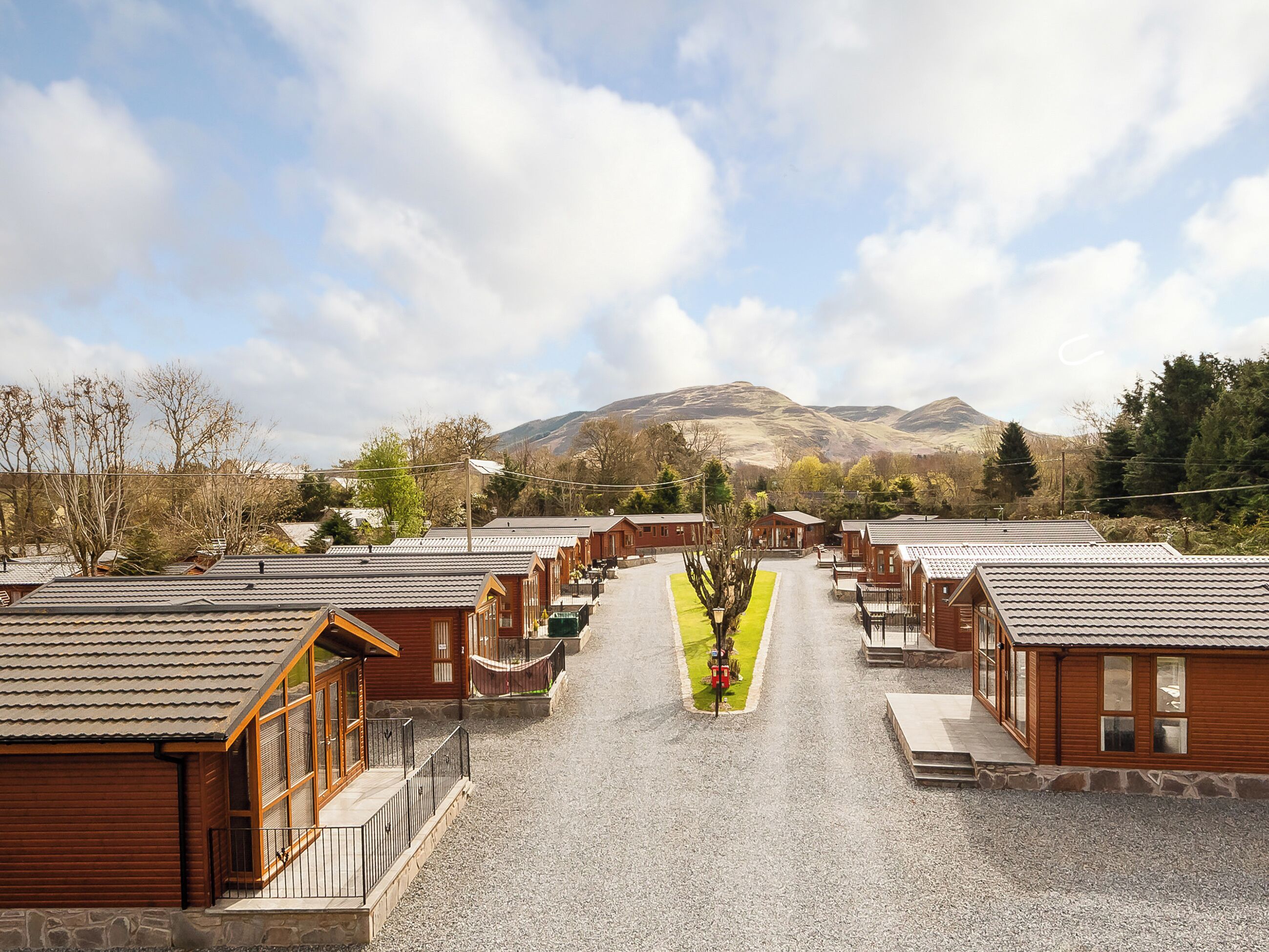 A row of wooden cabins with a gravel driveway leading to a central green area, surrounded by mountains and trees.
