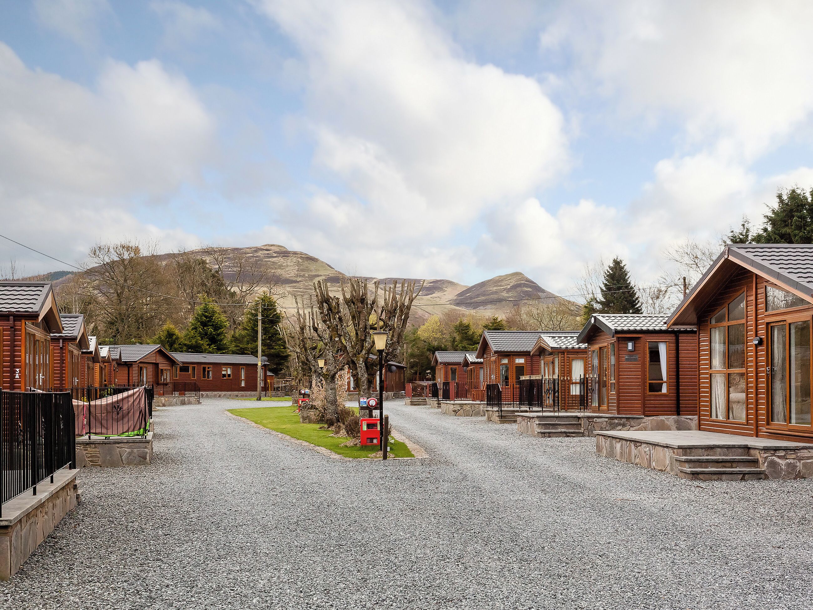 A row of wooden cabins with a gravel driveway, a red mailbox, and a mountain in the background.