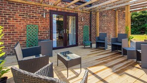Un patio couvert, aménagé avec des meubles en osier et une table en verre, jouxtant un bâtiment en briques surmonté d’une pergola en bois.
