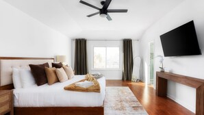 Un dormitorio con cama, ventilador de techo, televisión y ventana con cortinas.