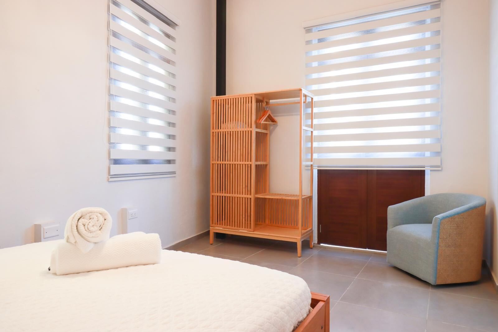 A bedroom with a bed, a wooden wardrobe, a chair, and a window with blinds.