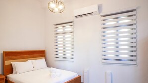 Un dormitorio con cama de madera, ropa de cama blanca, una lámpara de techo, equipo de aire acondicionado y dos ventanas con persianas.