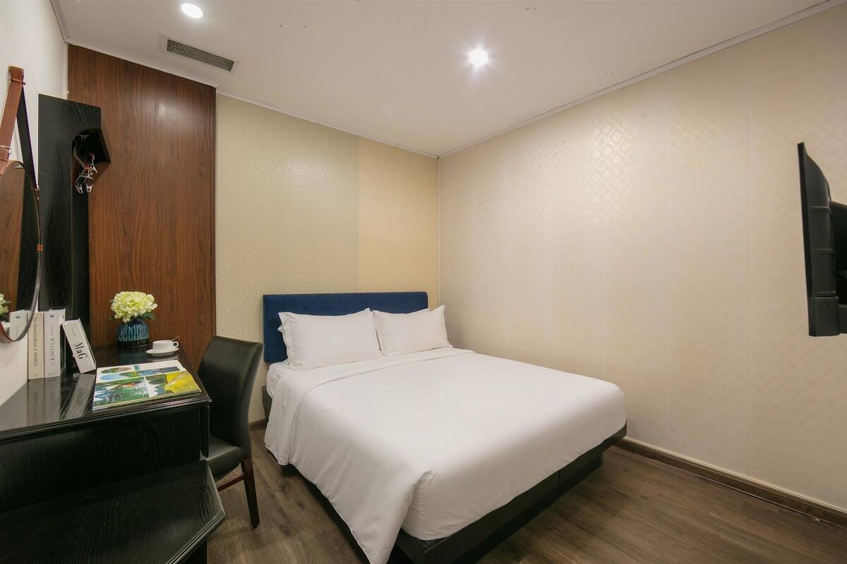 A hotel room with a bed, a desk with a chair, a small table with a vase of flowers, and a wall-mounted TV.