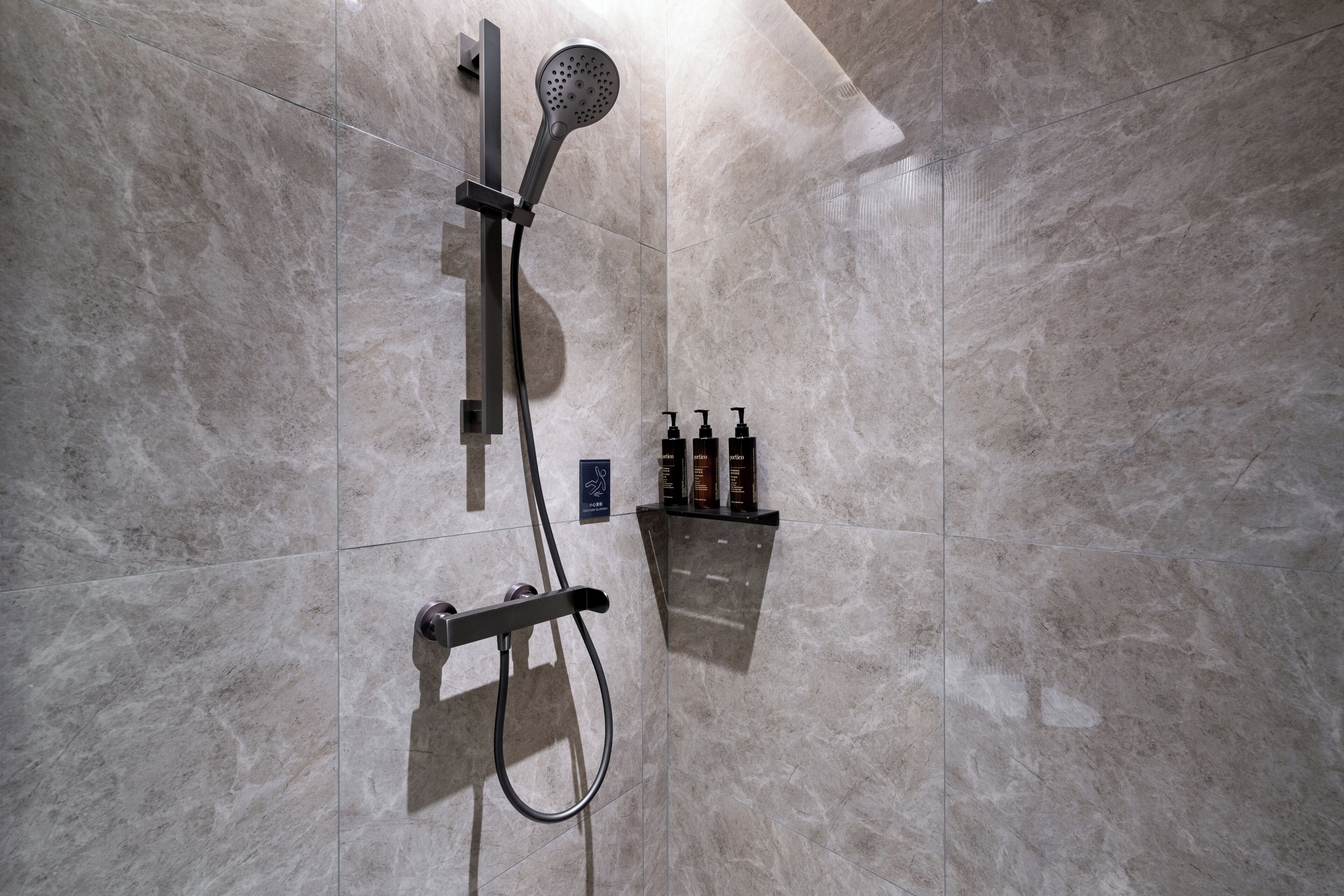 A shower with a handheld showerhead, a wall-mounted faucet, and a handheld shower hose. There are toiletries on a shelf below the shower.