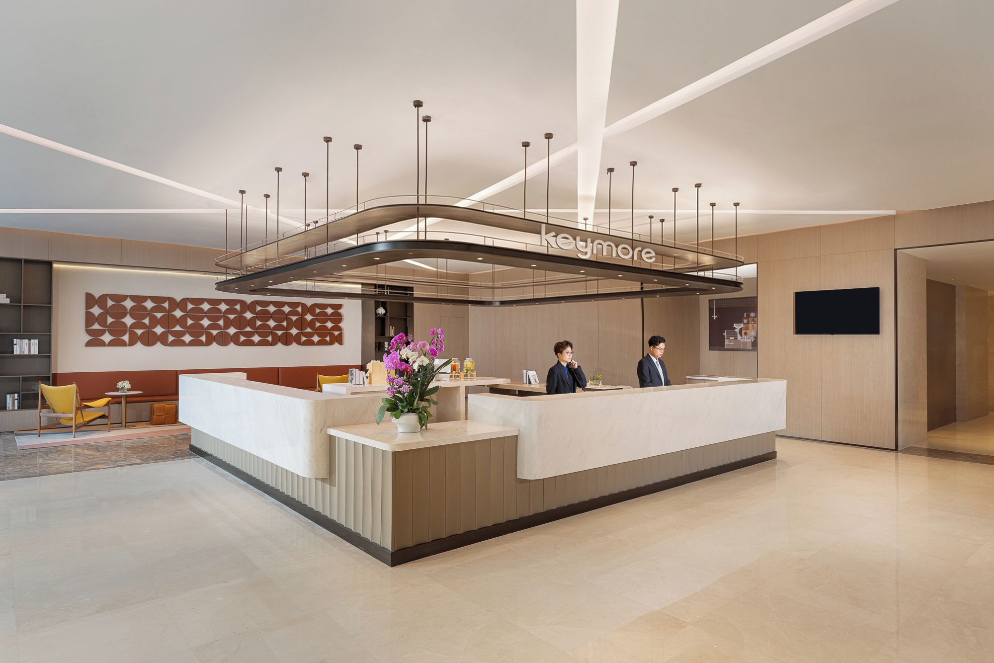 A modern reception desk with a glass partition, a seating area with a yellow chair, and a decorative wall with geometric patterns.
