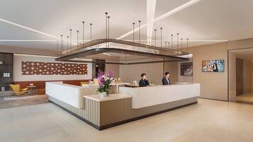 A modern hotel reception area with a curved reception desk, a large chandelier, and a wall-mounted digital display.