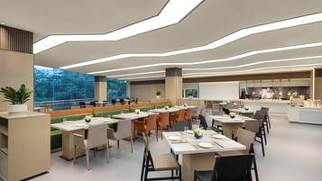 A modern dining area with multiple tables and chairs, a large window with a view of trees, and a kitchen area in the background.