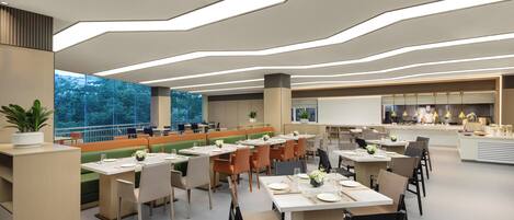 A modern dining area with multiple tables and chairs, a large window with a view of trees, and a kitchen area in the background.