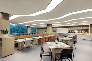 A modern dining area with multiple tables and chairs, a large window with a view of trees, and a kitchen area in the background. - Keymore Residence Guangzhou (Guangzhou)