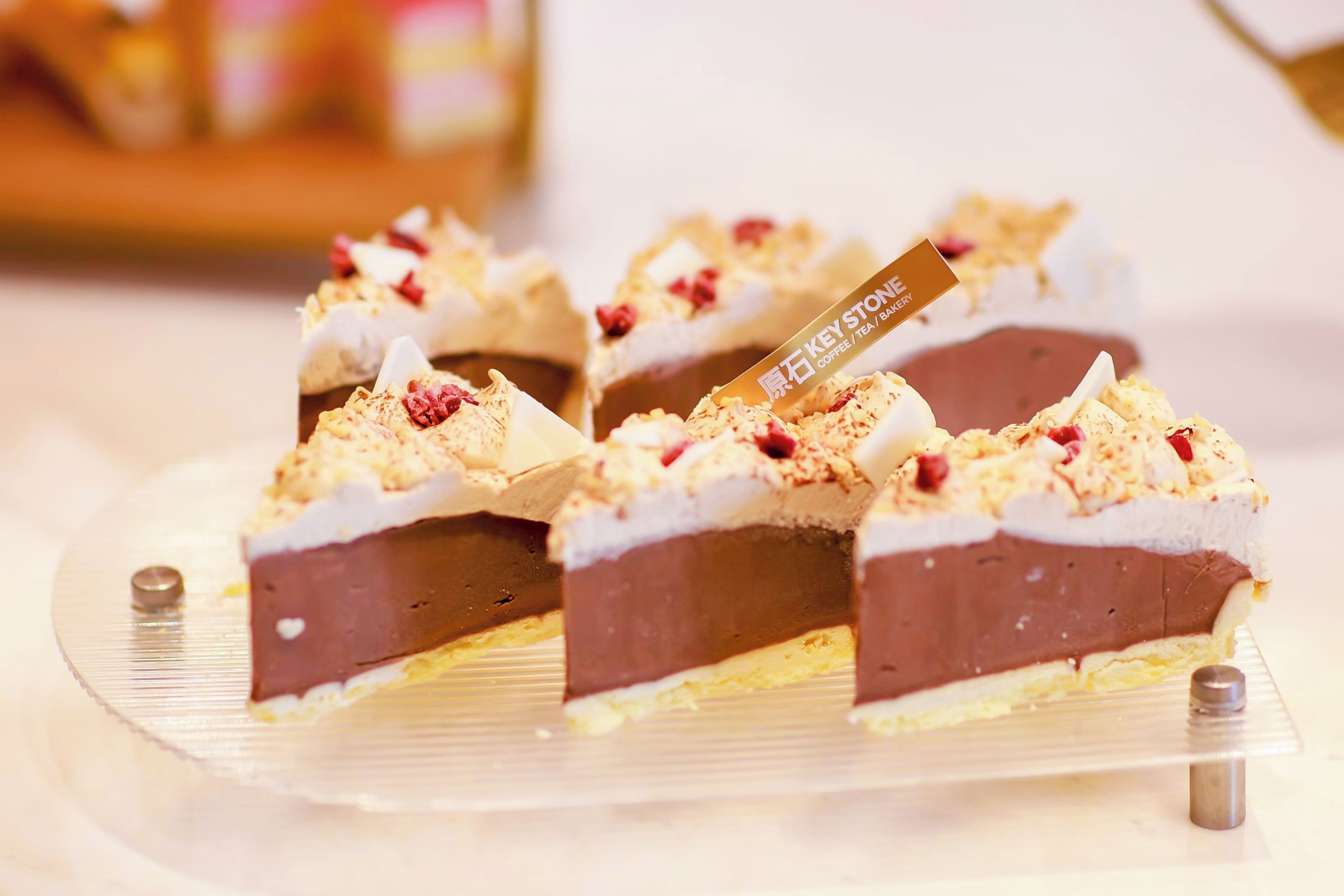 A dessert with a chocolate layer, white cream, and red fruit pieces, served on a clear glass plate with a metal stand.