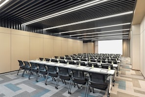 Une salle de conférence avec des rangées de tables et de chaises, un grand écran à l’avant et un tapis à motifs.