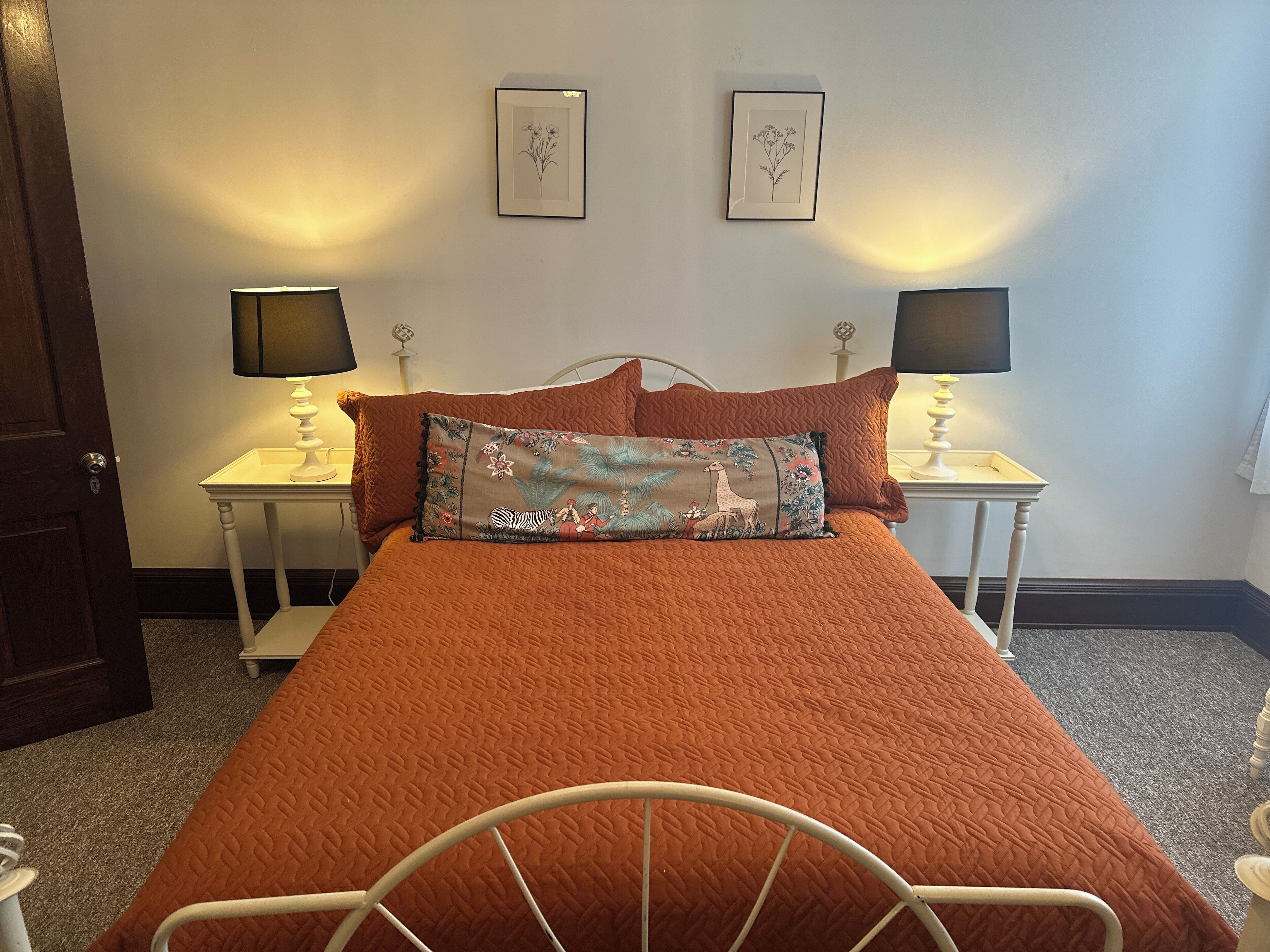 A neatly made bed with a brown quilt, two bedside tables with lamps, and two framed botanical prints on the wall.