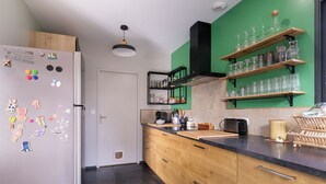 A modern kitchen with wooden cabinets, a black countertop, and a green backsplash. There are shelves with glassware and a coffee machine.