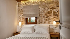 Een slaapkamer met een stenen muur, een raam met gordijn, twee bedlampen en een bed met een gestreept dekbed.
