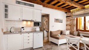 Een kleine keuken met witte kasten, een magnetron en een koffieapparaat. Er is een zithoek met een tafel en stoelen, en een bank met kussens. De kamer heeft een rustieke stenen muur en een houten plafondbalk.