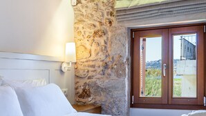 A bedroom with a stone wall, a bed with white linens, a nightstand with a lamp, and a view through a door.