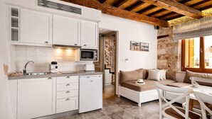 A compact kitchen with white cabinets, a microwave, and a small refrigerator. There is a dining area with a table and chairs, and a sofa with cushions. The room has a rustic stone wall and a wooden ceiling beam.