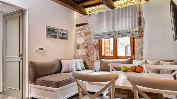 A cozy room with a stone wall, a wooden ceiling, a sofa, a dining table with chairs, and a window with blinds.