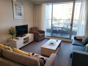 A modern living room with a flat-screen TV, striped armchair, sofa, and a balcony with outdoor furniture. - Glenelg Apartment near the Beach (Glenelg)
