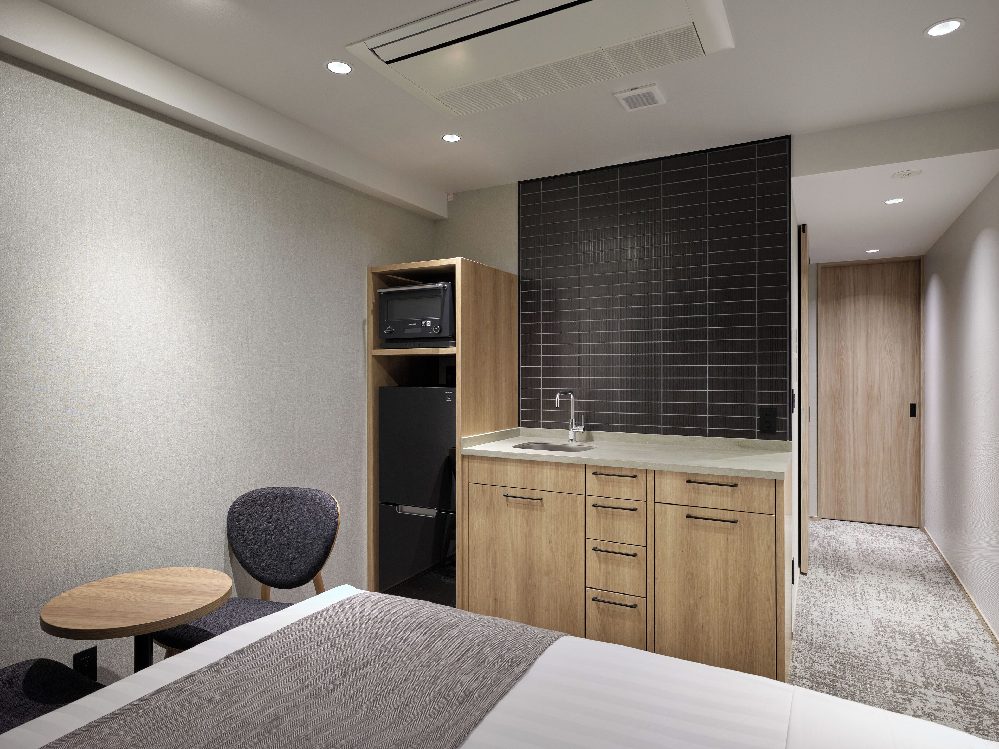 Una habitación de hotel moderna con cama, escritorio pequeño, silla, armario empotrado y kitchenette.