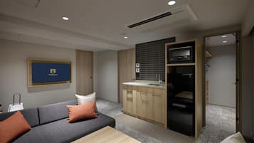 A modern hotel room with a grey sofa, a wooden coffee table, a kitchen area with a sink and stove, and a TV mounted on the wall.
