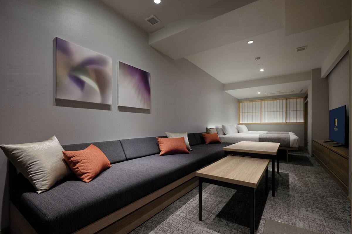 A modern living room with a dark grey sofa, a wooden coffee table, and a bed with a white headboard. There are abstract paintings on the wall and a television on the right.