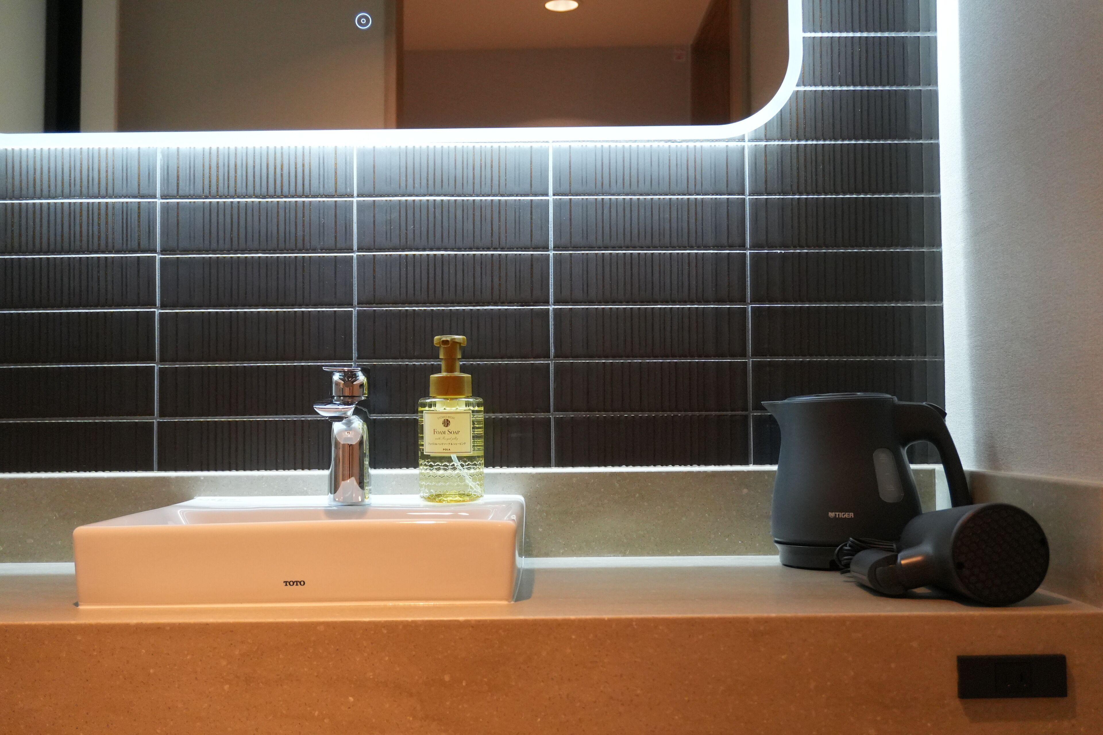 A bathroom with a sink, a bottle of hand soap, a black kettle, and a hairdryer on the countertop.