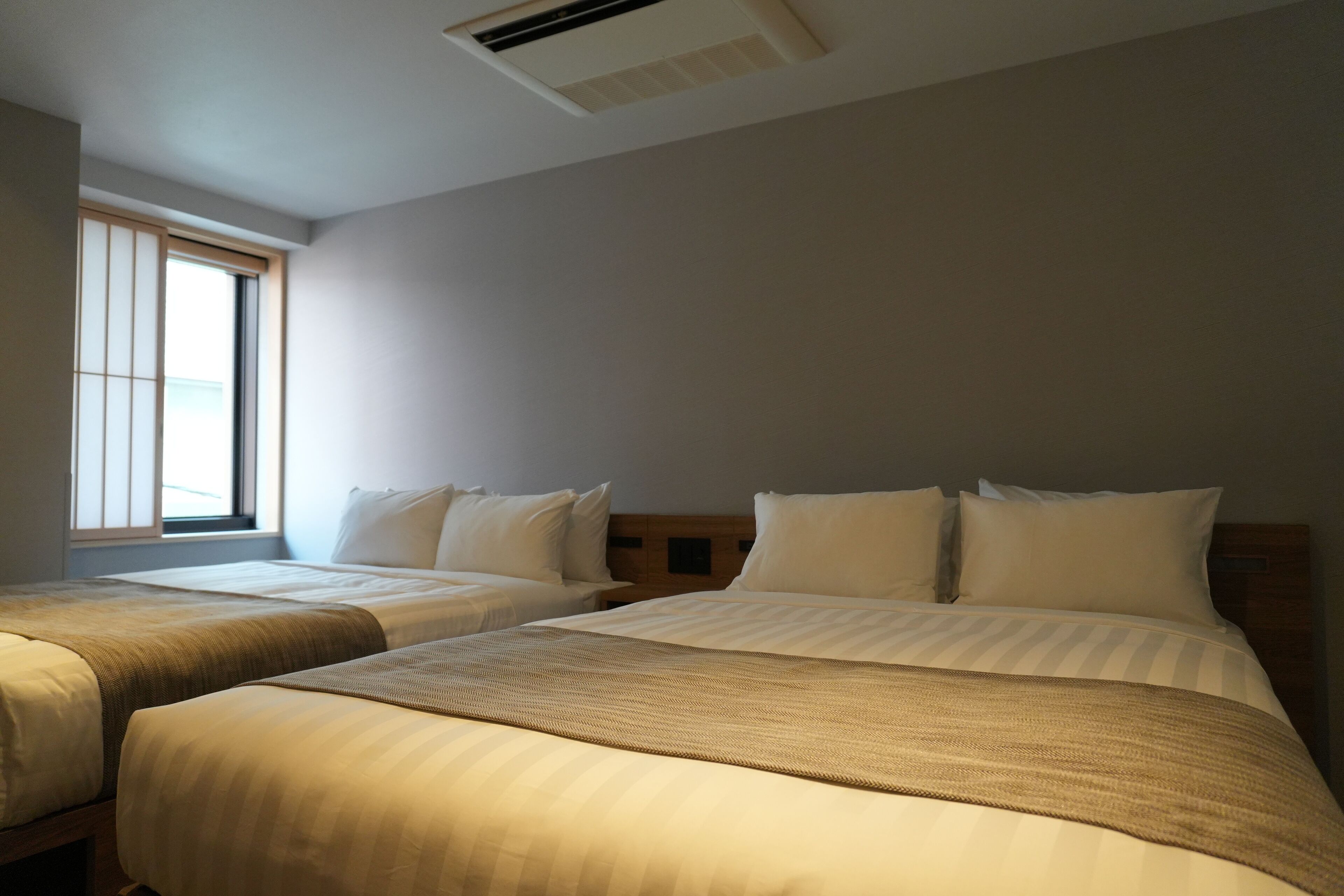 A hotel room with two single beds, a large window, and a ceiling air vent.