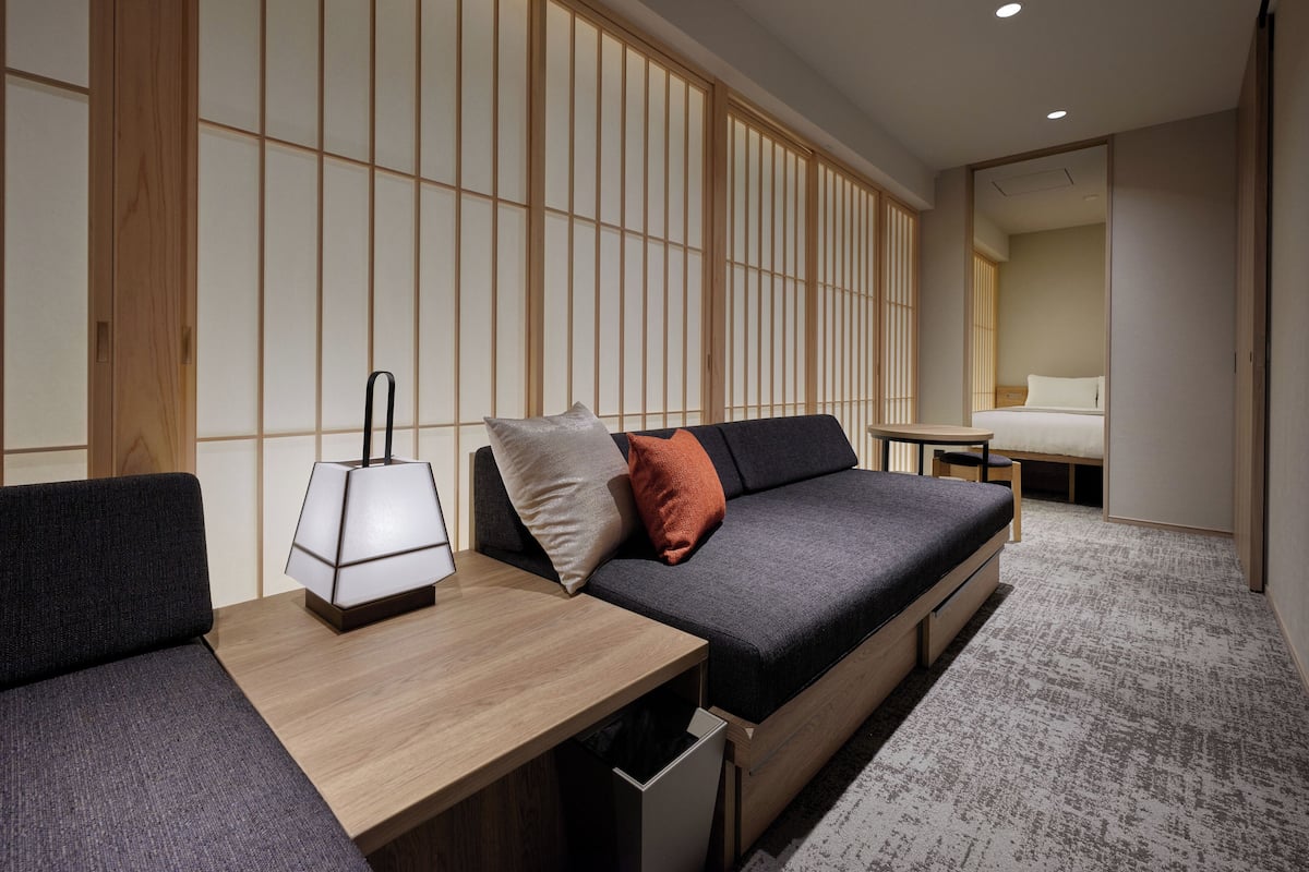 A modern room with a sofa, a small table with a lamp, and a bed in the background.