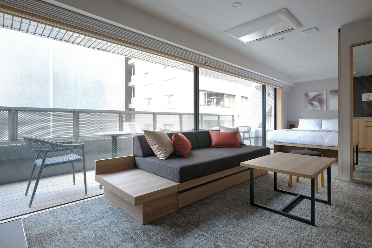 A modern hotel room with a sofa, a small table, a bed, and a balcony with a chair.