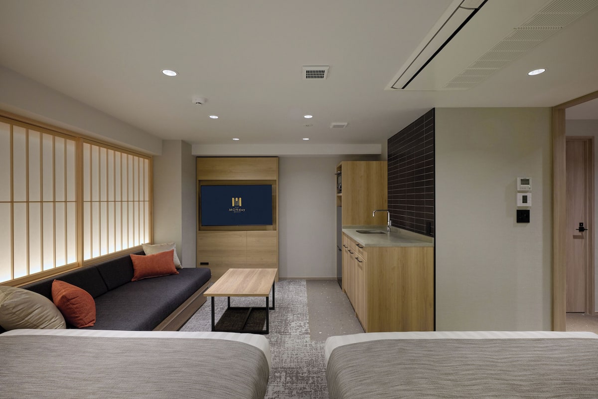 A modern hotel room with a sofa, two beds, a small table, a kitchenette, and a TV mounted on the wall.