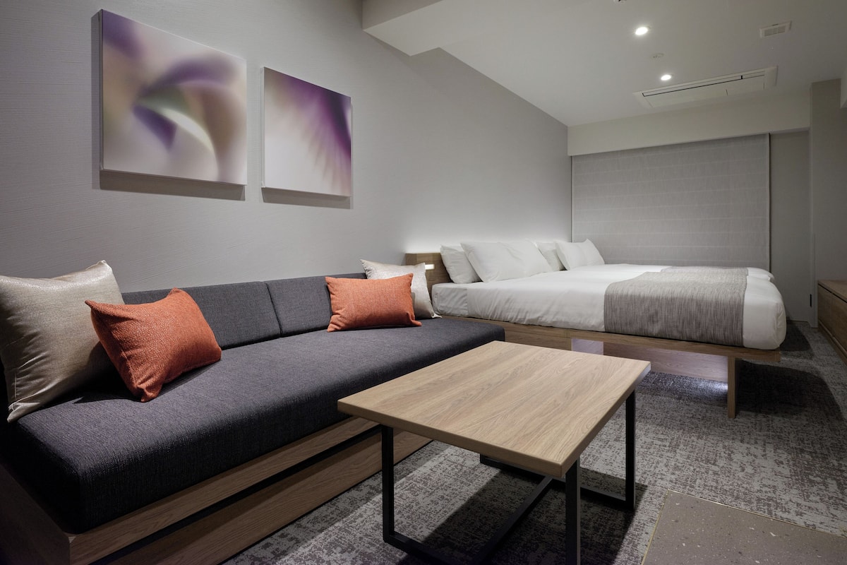 A modern hotel room with a grey sofa, a wooden coffee table, a bed with white bedding, and abstract wall art.
