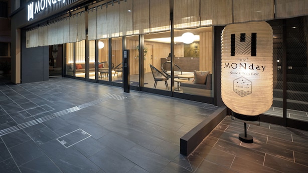 L’extérieur d’un hôtel moderne, avec une grande enseigne indiquant « LUNDI », de larges fenêtres en verre et un espace de réception visible à l’intérieur.