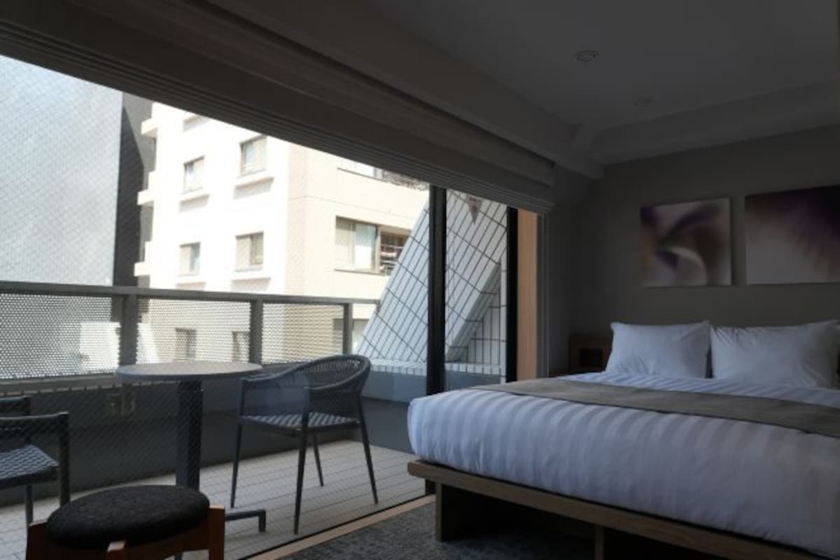 Habitación de hotel con cama, una mesita y una silla en un balcón que ofrece vistas a edificios.