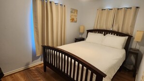 A bedroom with a wooden bed, beige curtains, a ceiling fan, and a nightstand with a lamp.