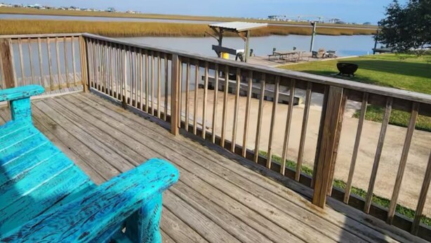 A wooden deck with a railing overlooking a body of water, a covered picnic area, and a grassy area with a tree.